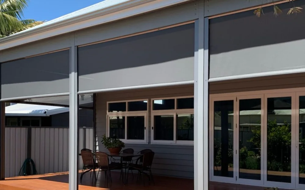 Charcoal Zipscreen blinds half lowered across the verandah of a weatherboard house, above a timber deck set with an outdoor dining table