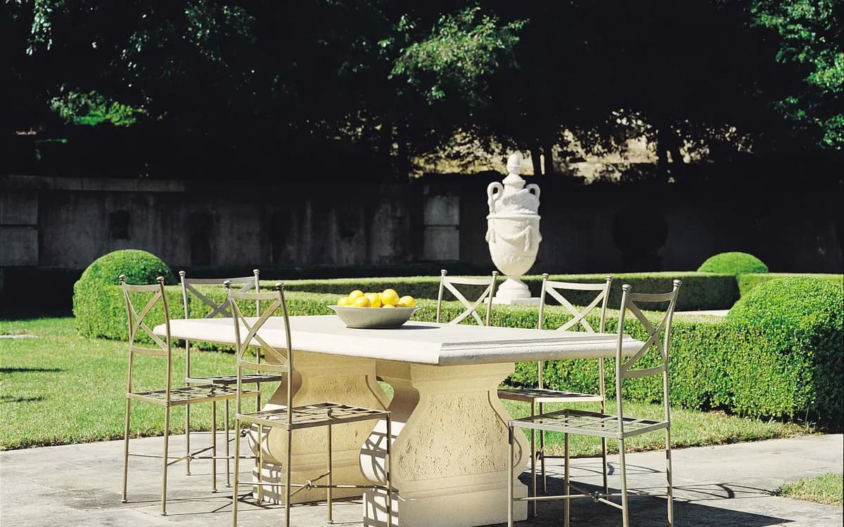A pale stone dining table with metal chairs on a garden terrace, a white urn and clipped hedges behind