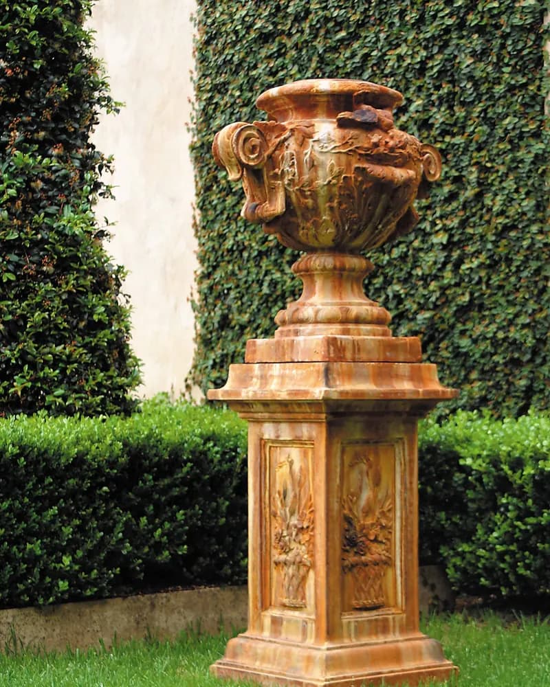 An ornate rust finished urn on a carved pedestal, against a clipped hedge