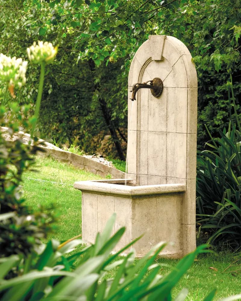 A stone wall fountain with a bronze tap, standing in a green garden